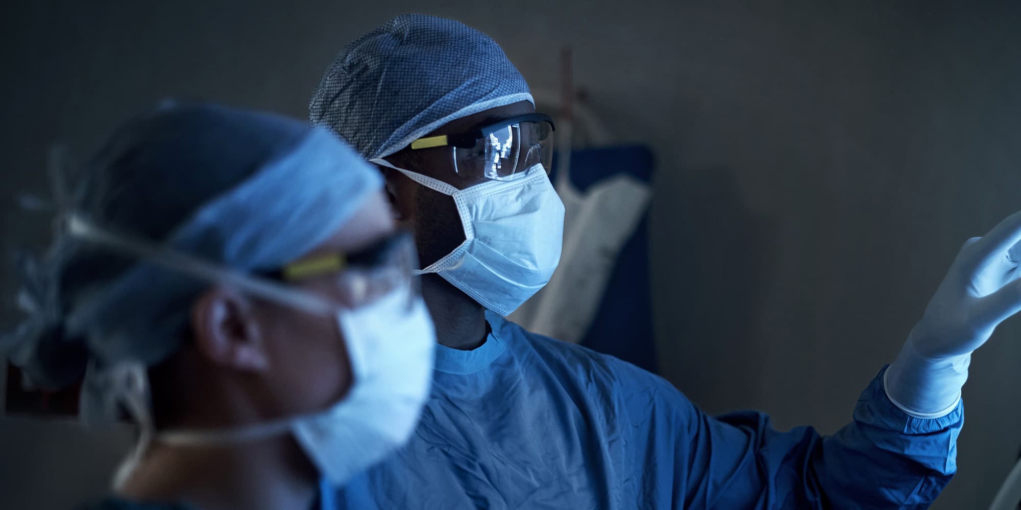 HSI_SURGERY_WEB_VISUAL_2000x1000 Surgeons in masks and protective gear working in a dim operating room
