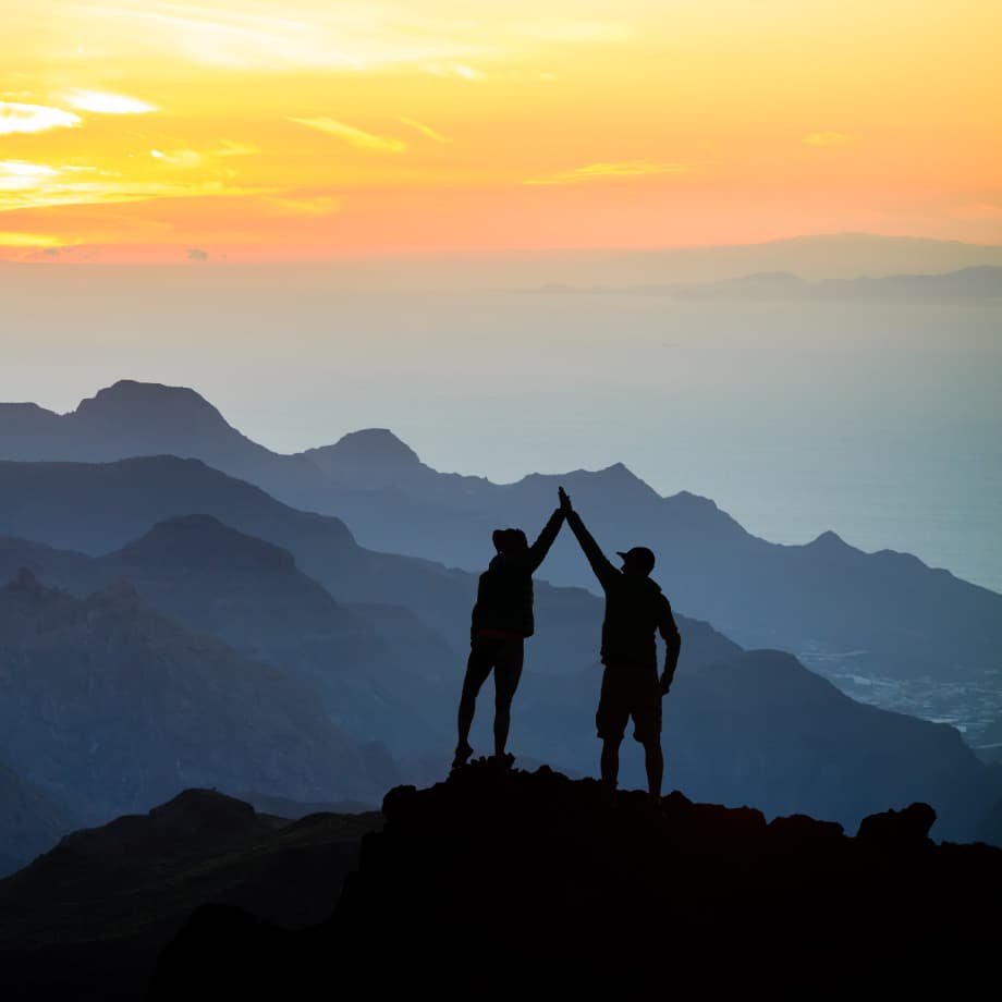 Design-and-IP-partners Two people silhouetted on a mountain peak raise their hands against a sunrise sky