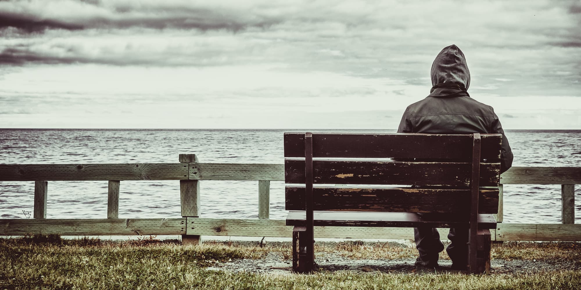 03_kunnen_we_gelukkig_zijn_meten_1 A hooded person sits alone on a bench facing the sea under a cloudy sky