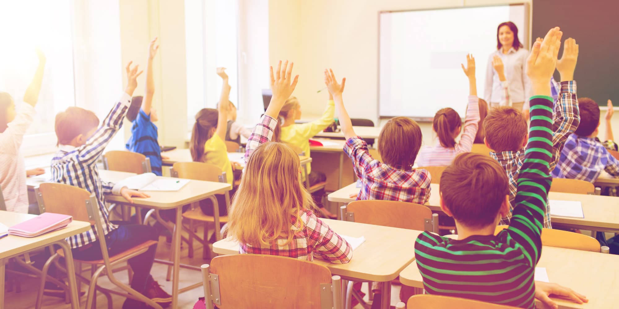 07_DCAP_spinoff_1 Primary school classroom with children raising their hands while a teacher stands at the front