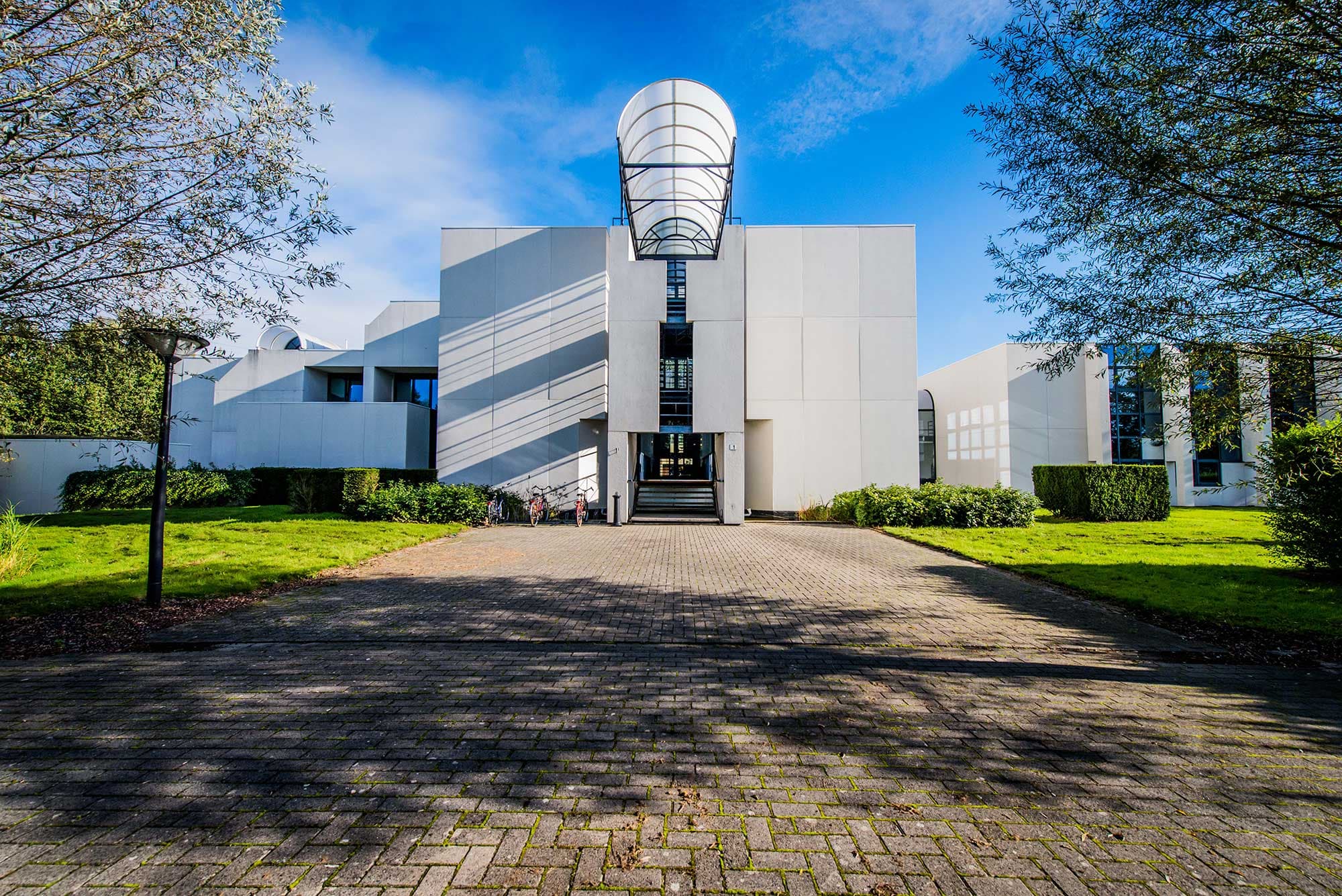 1_building Modern white research building with a curved glass entrance canopy, framed by trees and green lawn.