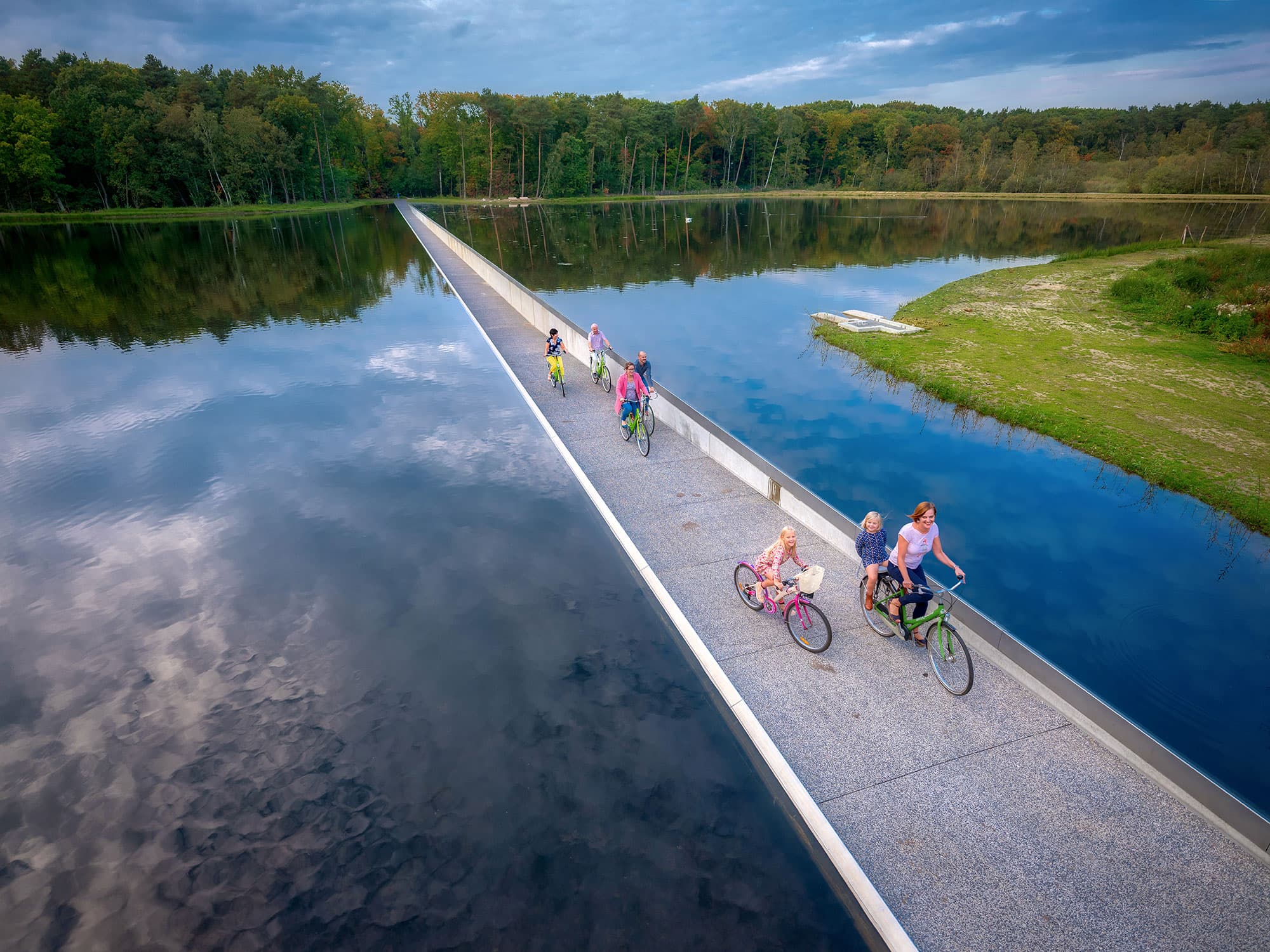 2017_Toerisme_Limburg_Cycling_through_Water_Family_Picture_0 Cyclists riding along a narrow bridge over a lake, surrounded by trees and open water.