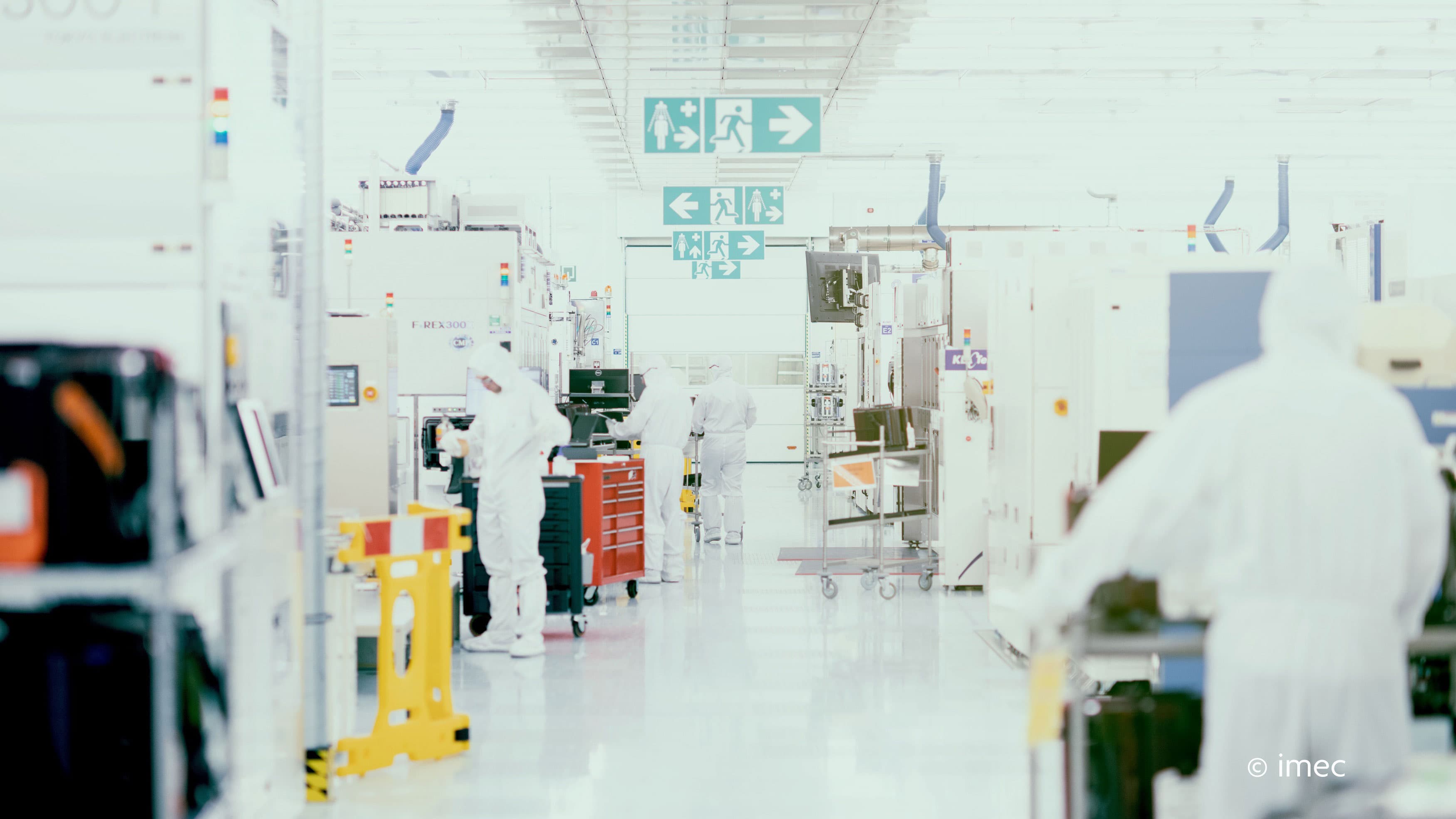 2019_imec_cleanroom04_header Cleanroom workers in white suits move between semiconductor equipment in imec’s lab