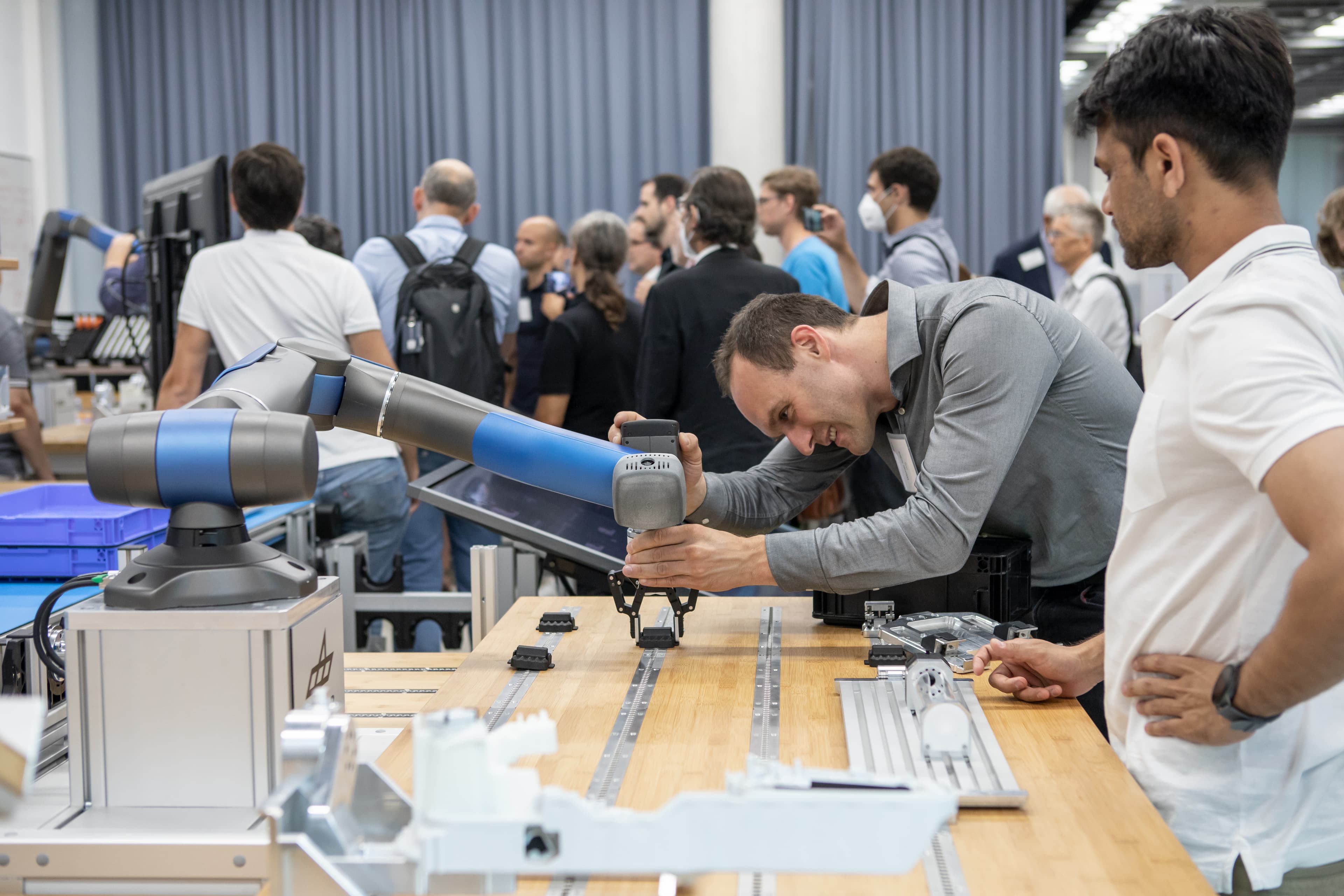 2022_euROBIN_0J4A0749 Researchers demonstrate a robotic arm at a robotics showcase, with attendees watching in the background.