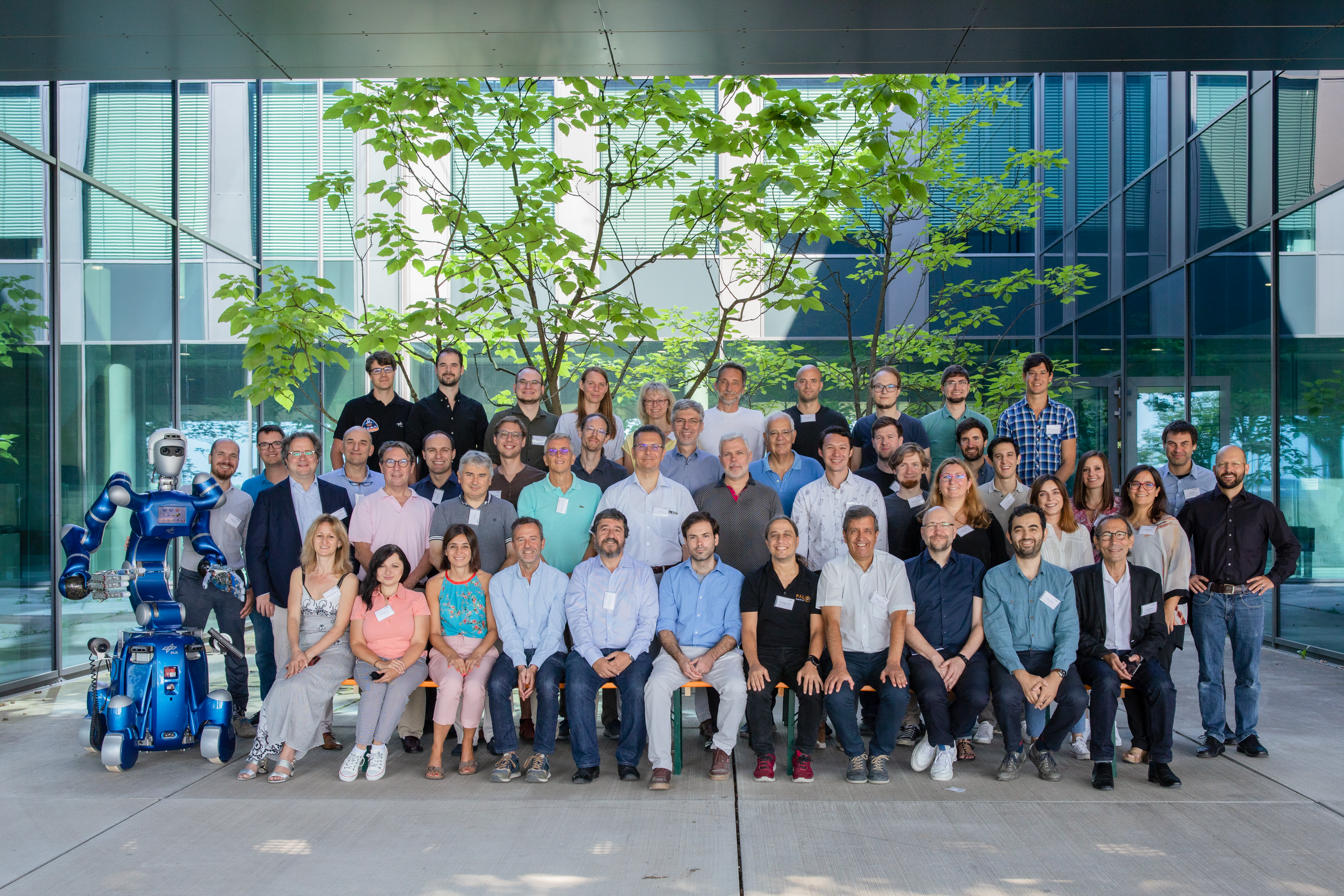 2022_euROBIN_Group%20Photo_0J4A0970 Group photo of EuROBIN participants with a blue humanoid robot at left, standing outside a modern building.