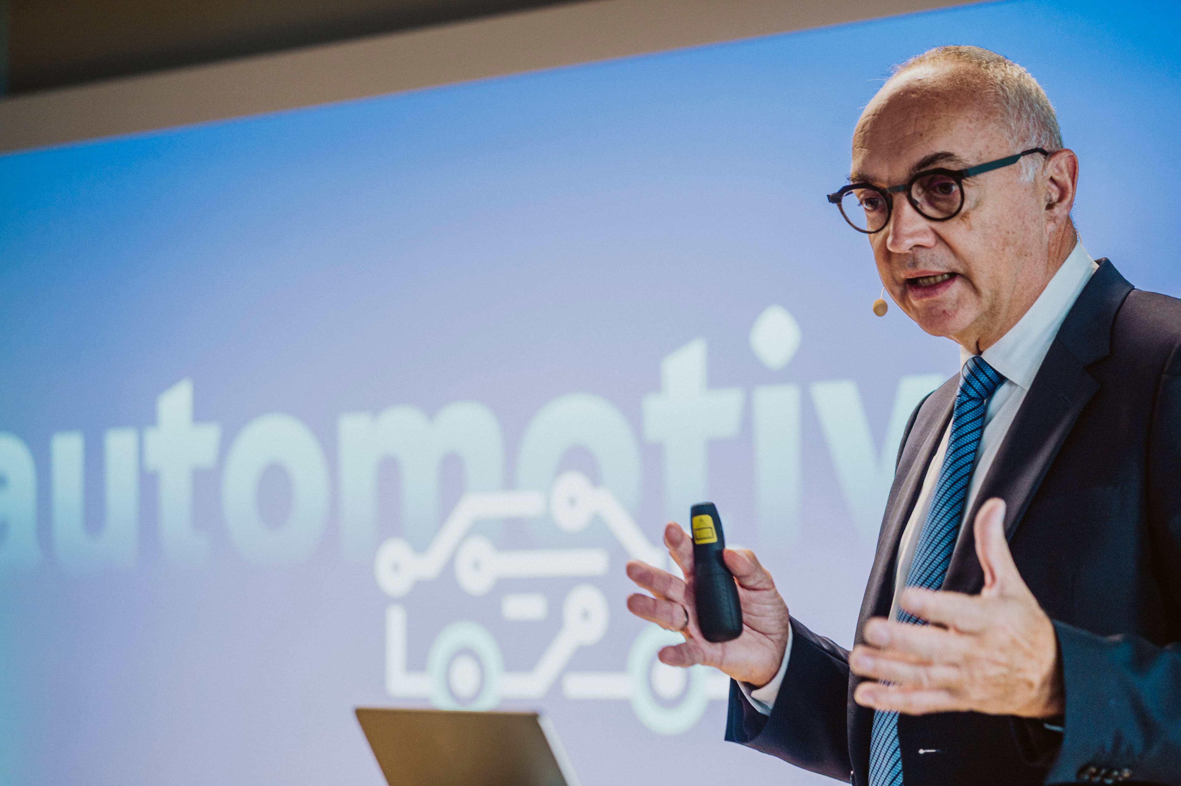 2025_6th_Automotive_Chiplet_Forum_imec_16 Speaker in a suit giving a presentation with an automotive chiplet graphic on the screen behind him.