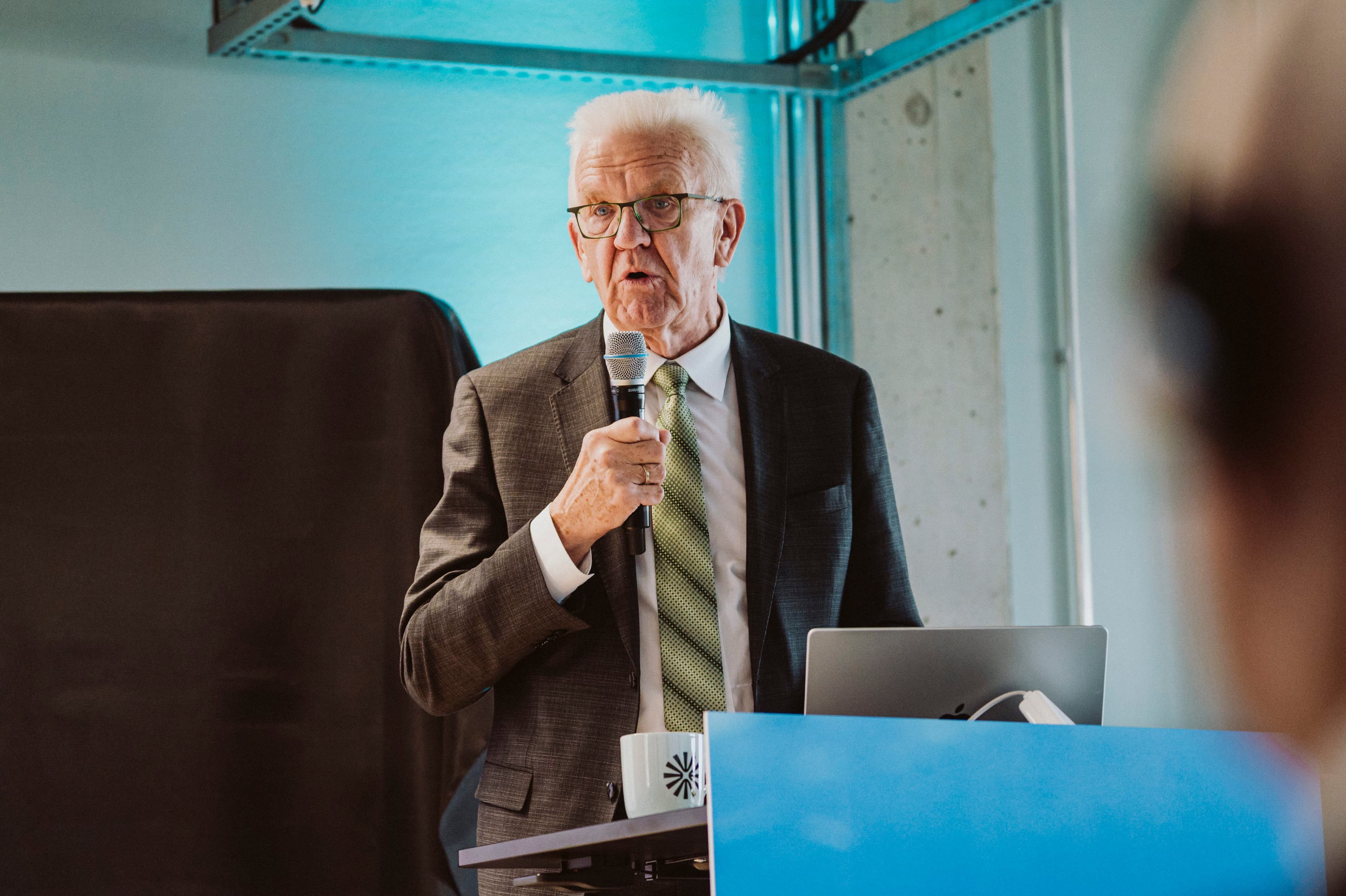 2025_6th_Automotive_Chiplet_Forum_imec_22 An older man in a suit speaks into a microphone at a podium during a presentation.