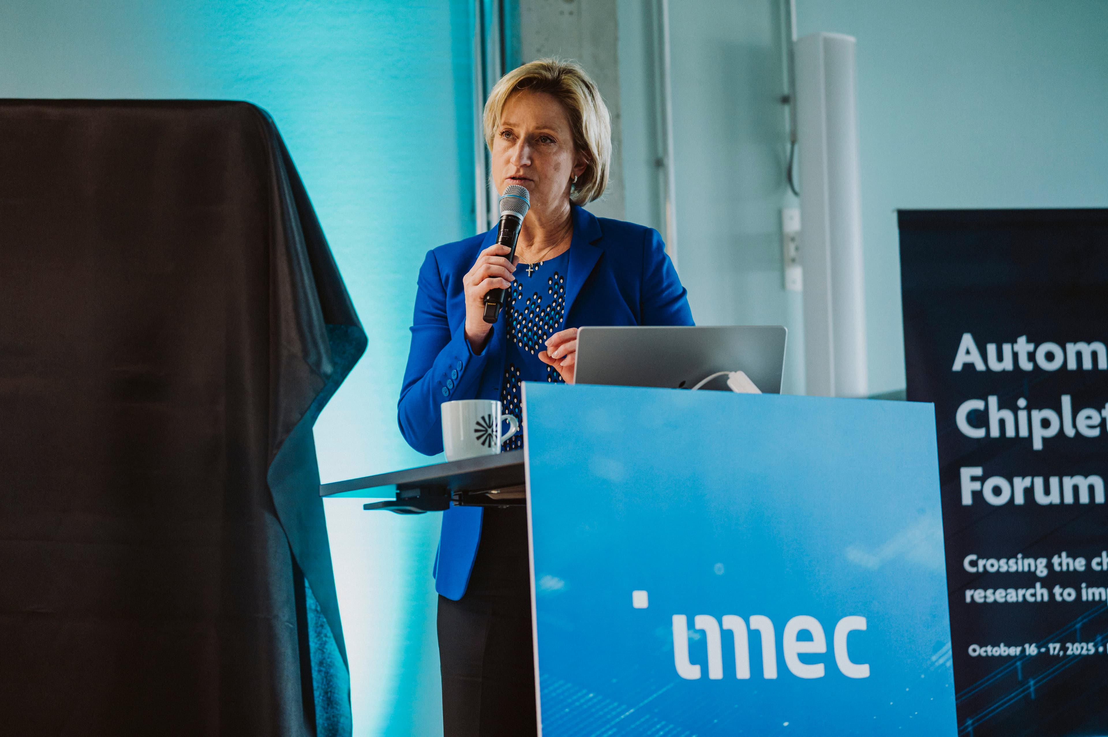 2025_6th_Automotive_Chiplet_Forum_imec_25 Speaker in a blue blazer giving a presentation at an imec automotive chiplet forum podium.