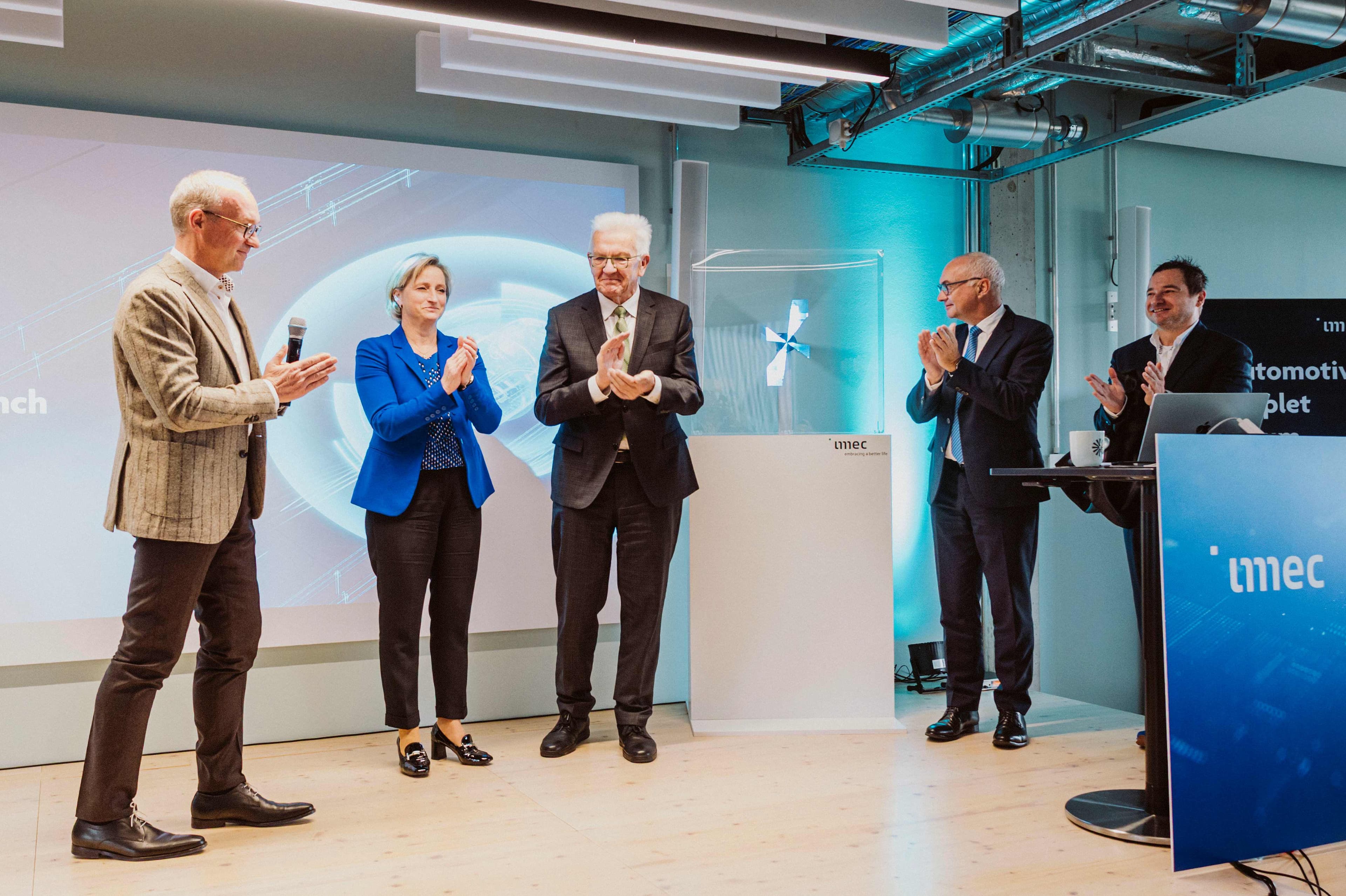 2025_6th_Automotive_Chiplet_Forum_imec_36 Speakers and guests applaud on a stage during an imec automotive chiplet event.