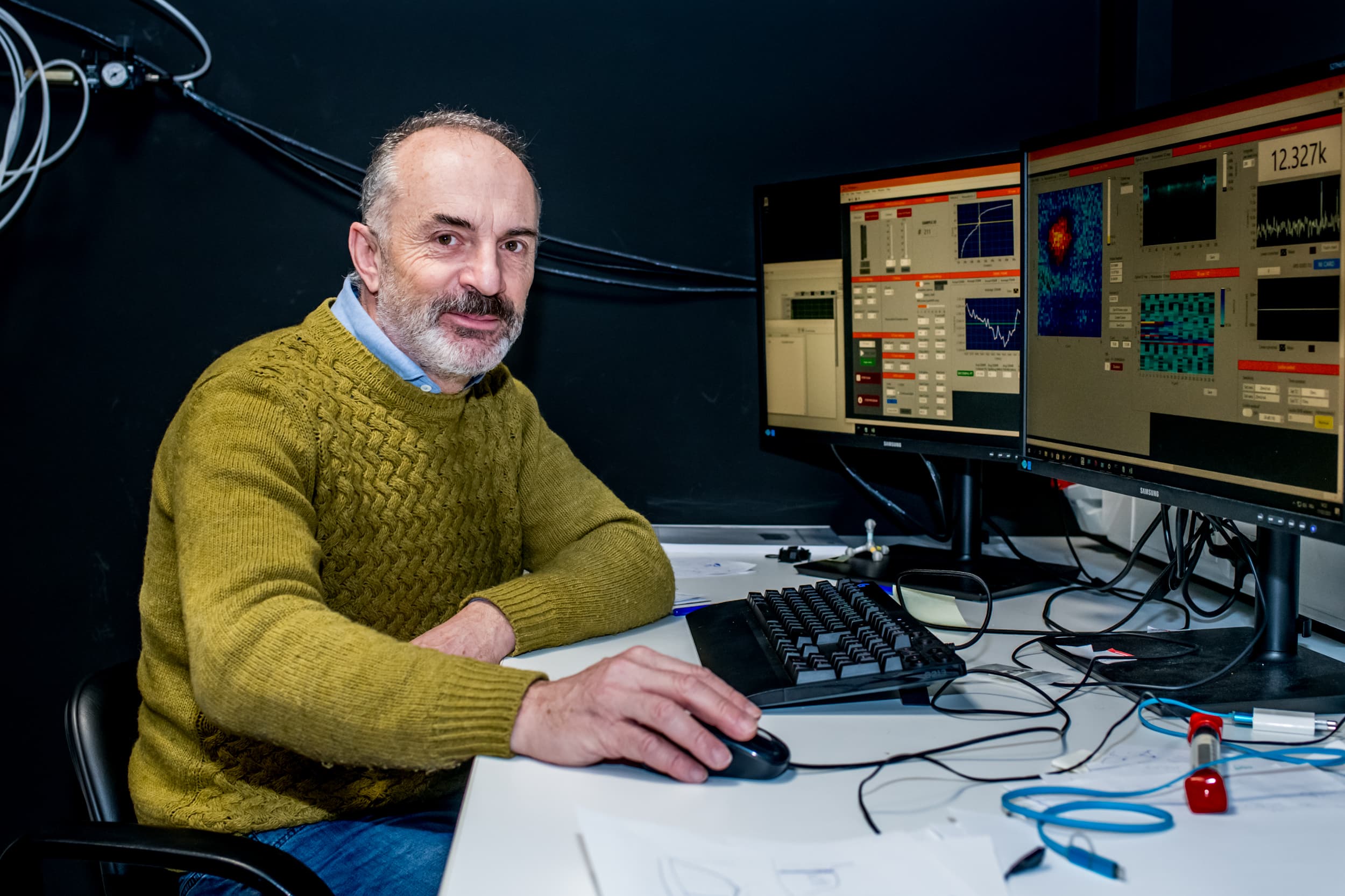 Man at lab desk with measurement monitors Milos Nesladek seated at a lab desk with dual monitors showing measurement graphs and a data interface