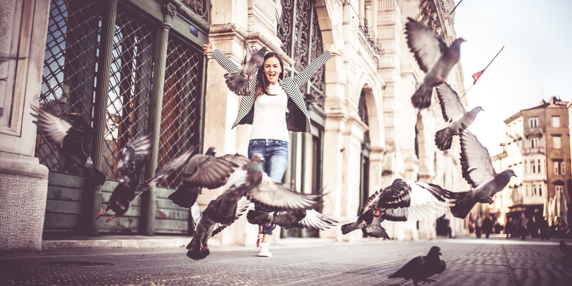 5_ai-voor-meer-avontur-in-de-stad_1 Woman with arms raised on a city street as pigeons fly around her near historic buildings.