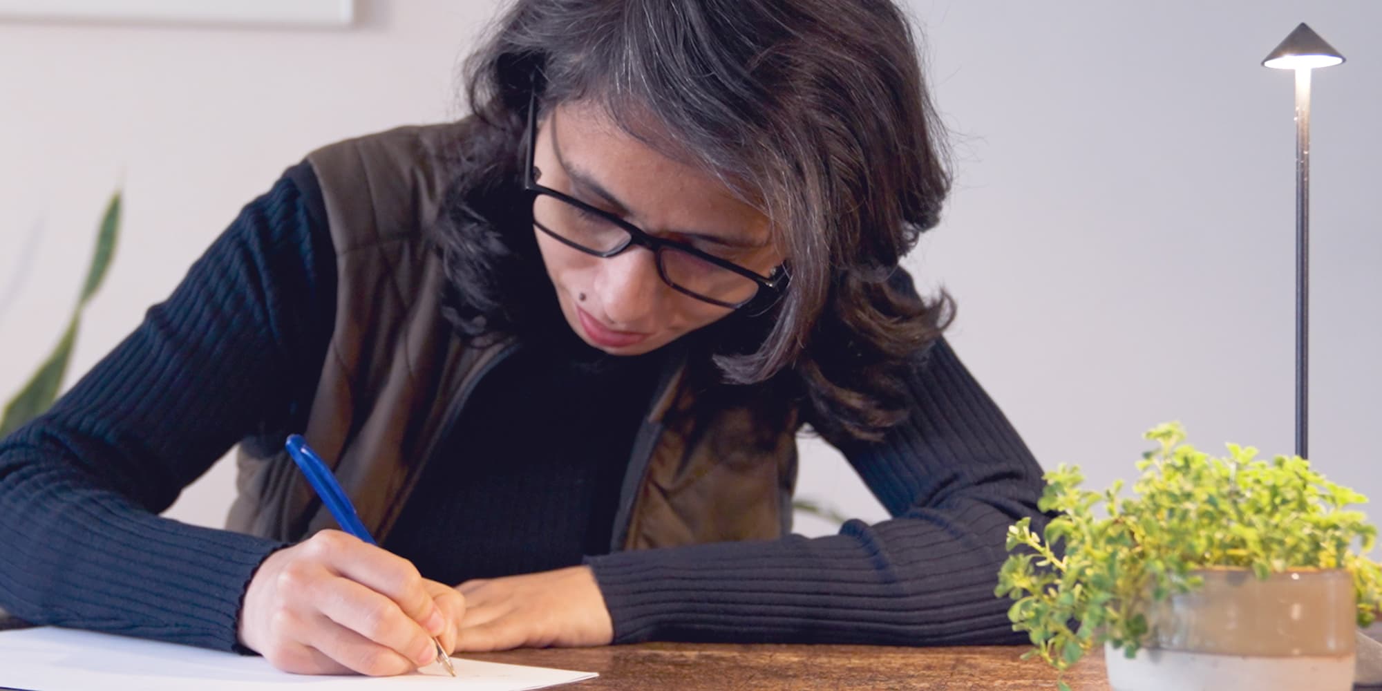 Aarti-Rathi-1 Aarti Rathi writing at a desk beside a small potted plant, looking down in concentration