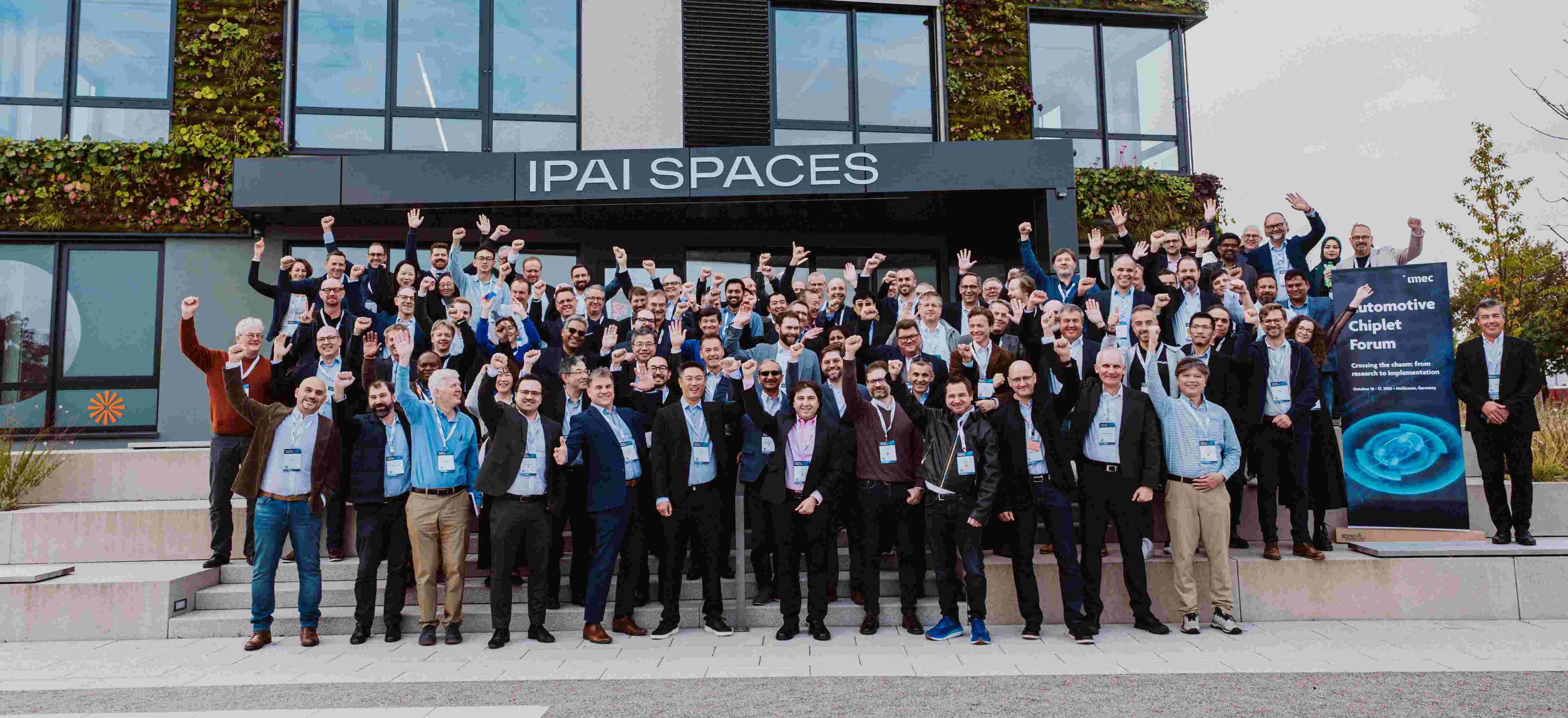 Group photo at Automotive Chiplet Forum conference Large group of conference attendees posing with raised hands outside IPAI Spaces beside an Automotive Chiplet Forum banner