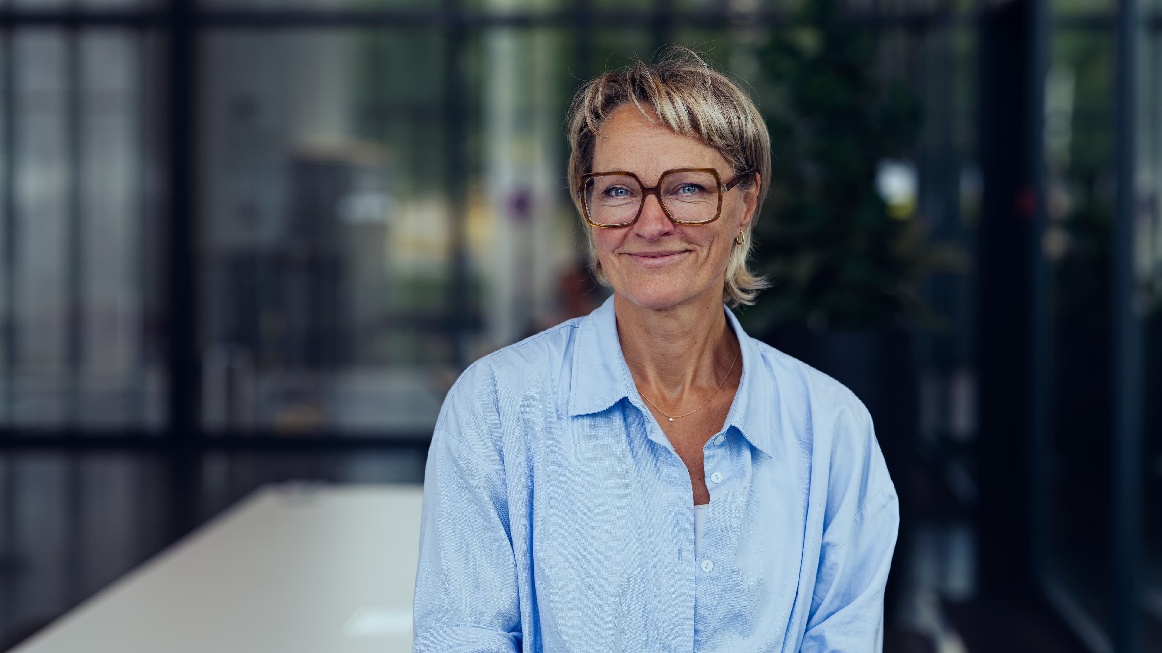 Annelies-Daenen-001 Portrait of Annelies Daenen wearing glasses and a light blue shirt, standing in front of a blurred background