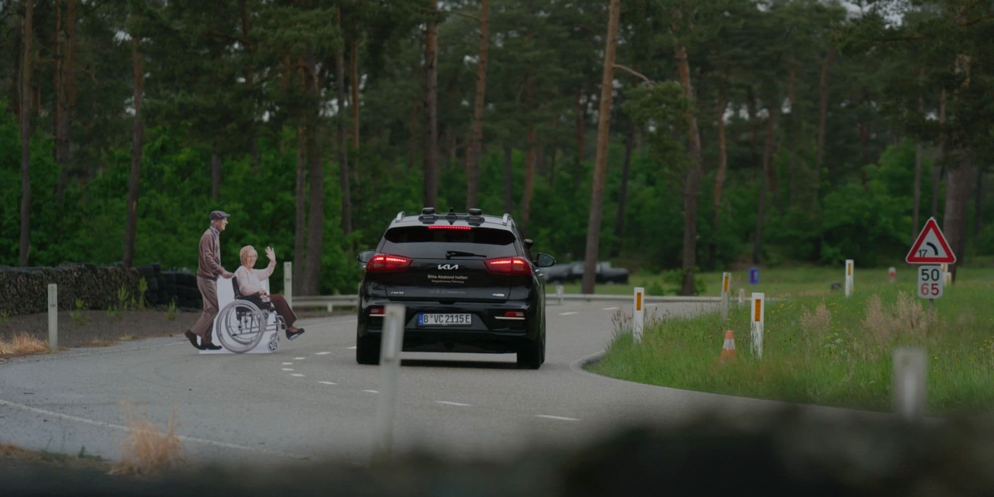 Autonome-auto-PR Zwarte autonome auto die over een bosweg rijdt terwijl een man een vrouw in een rolstoel ernaast duwt