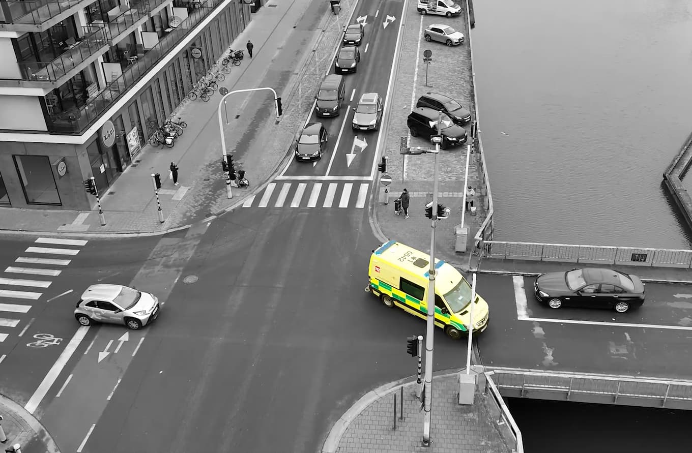 Beeld-ambu_0 Ambulance nadert een kruispunt naast een kanaal, met verkeerslichten en wachtende auto's in de buurt