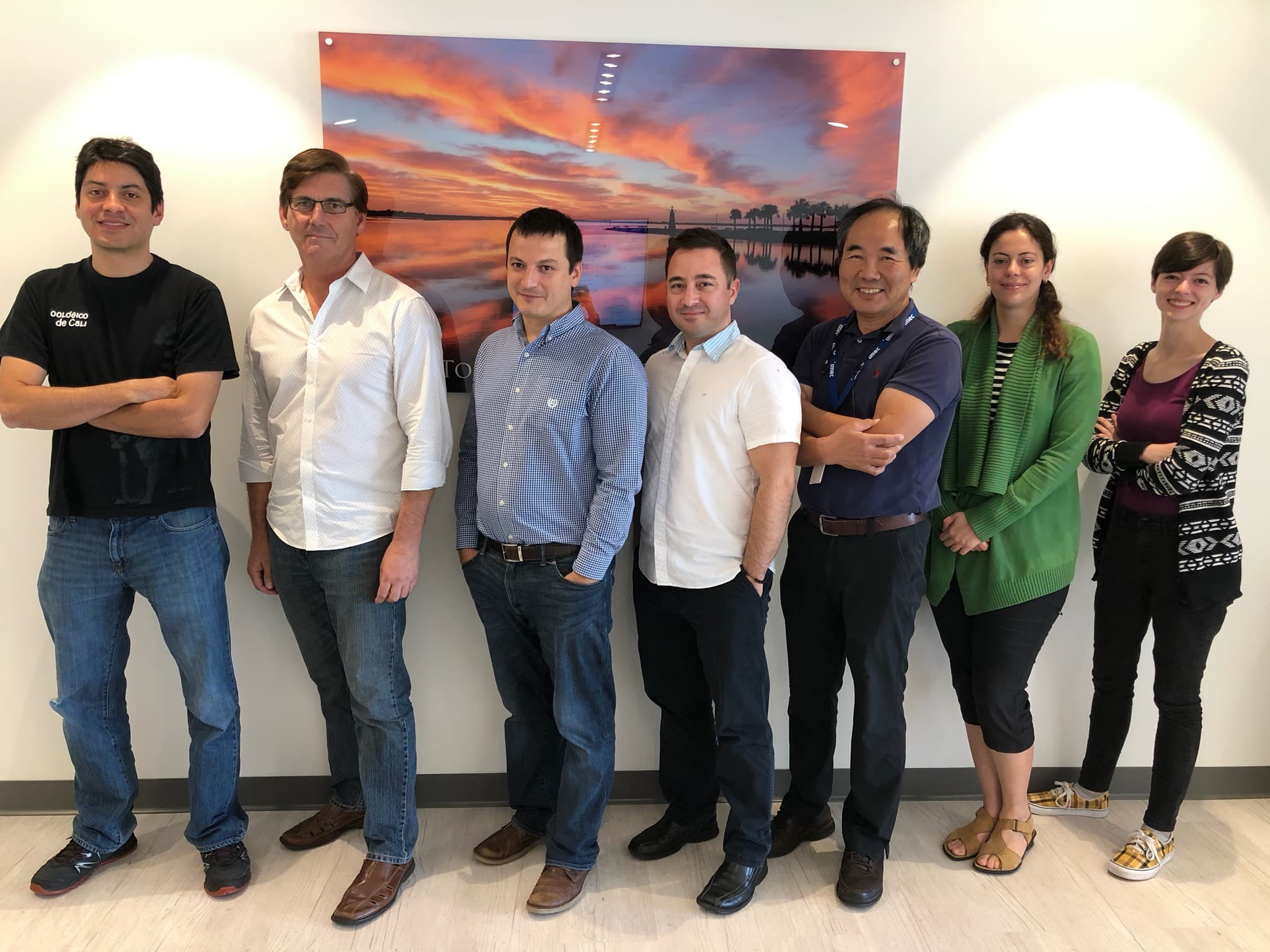 bio_imagingflorida_1 Seven researchers standing in a row indoors beneath a sunset wall print