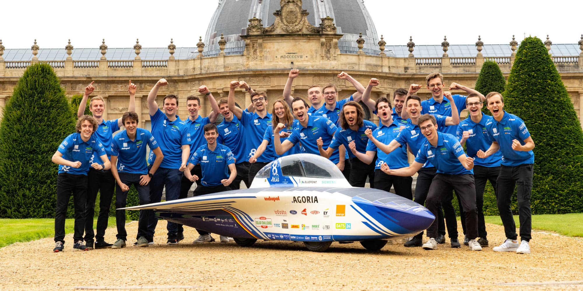 Bluepoint-atlas Team in blue shirts posing behind a solar-powered racing car, cheering in front of a historic building