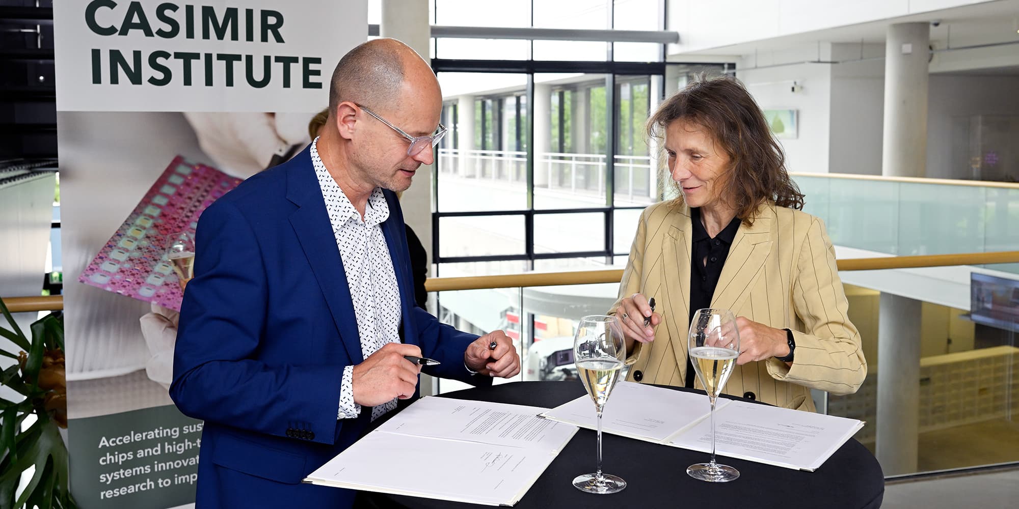 BvOF_2026_0608_0108-collaboration-signing-IMEC-and-TUe Two people signing a collaboration agreement beside a Casimir Institute banner, with champagne glasses on the table.