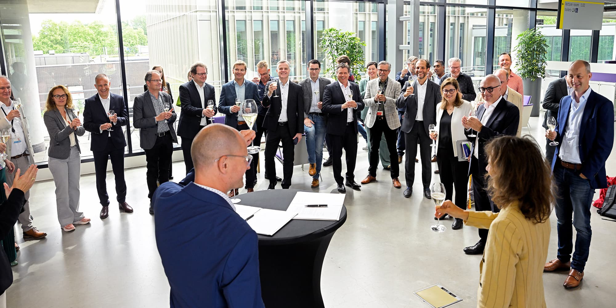 BvOF_2026_0608_0148-collaboration-signing-IMEC-and-TUe Group of people raising glasses at a signing reception in a bright atrium