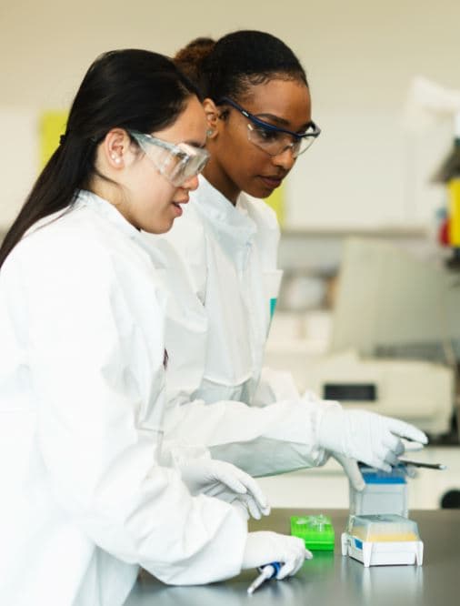 Capture_8 Two JHU students in lab coats and safety goggles work together at a bench in imec’s Life Sciences department.