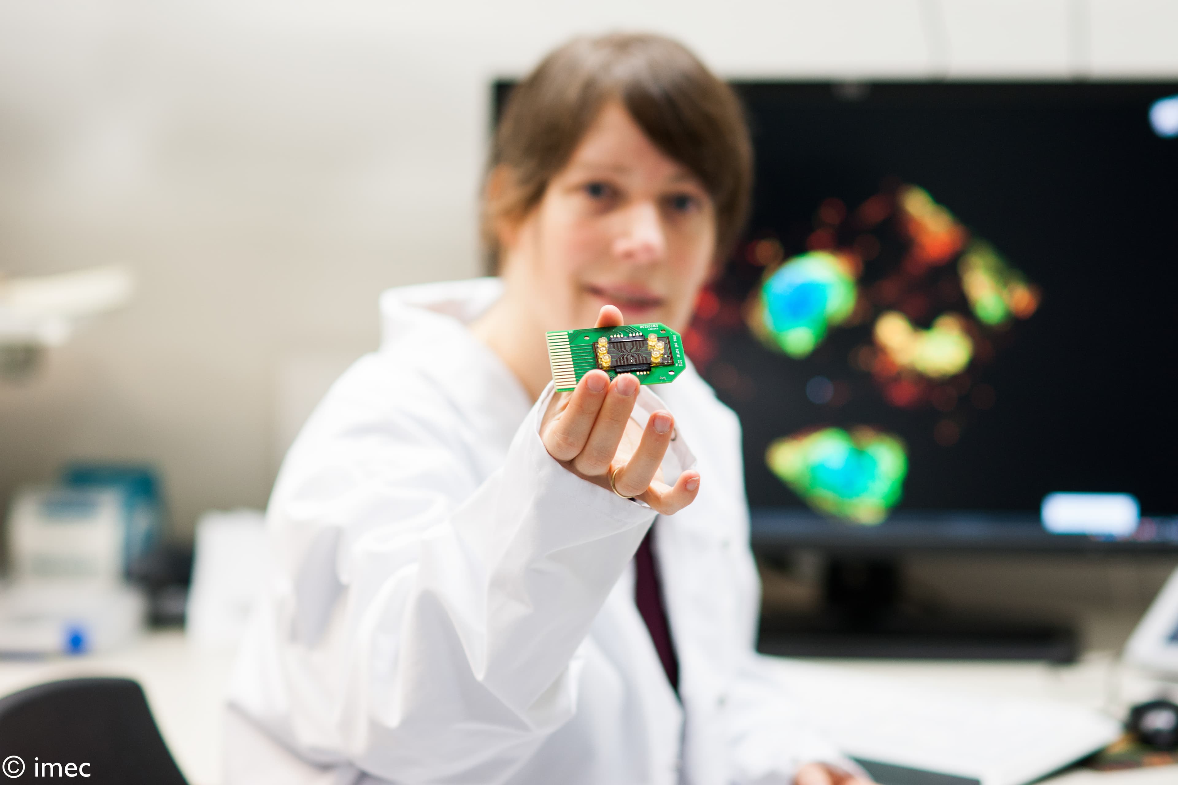 Cell_sorter_Liesbet_0 Researcher in a lab coat holding up a small cell sorter chip, with a colorful microscope image in the background.