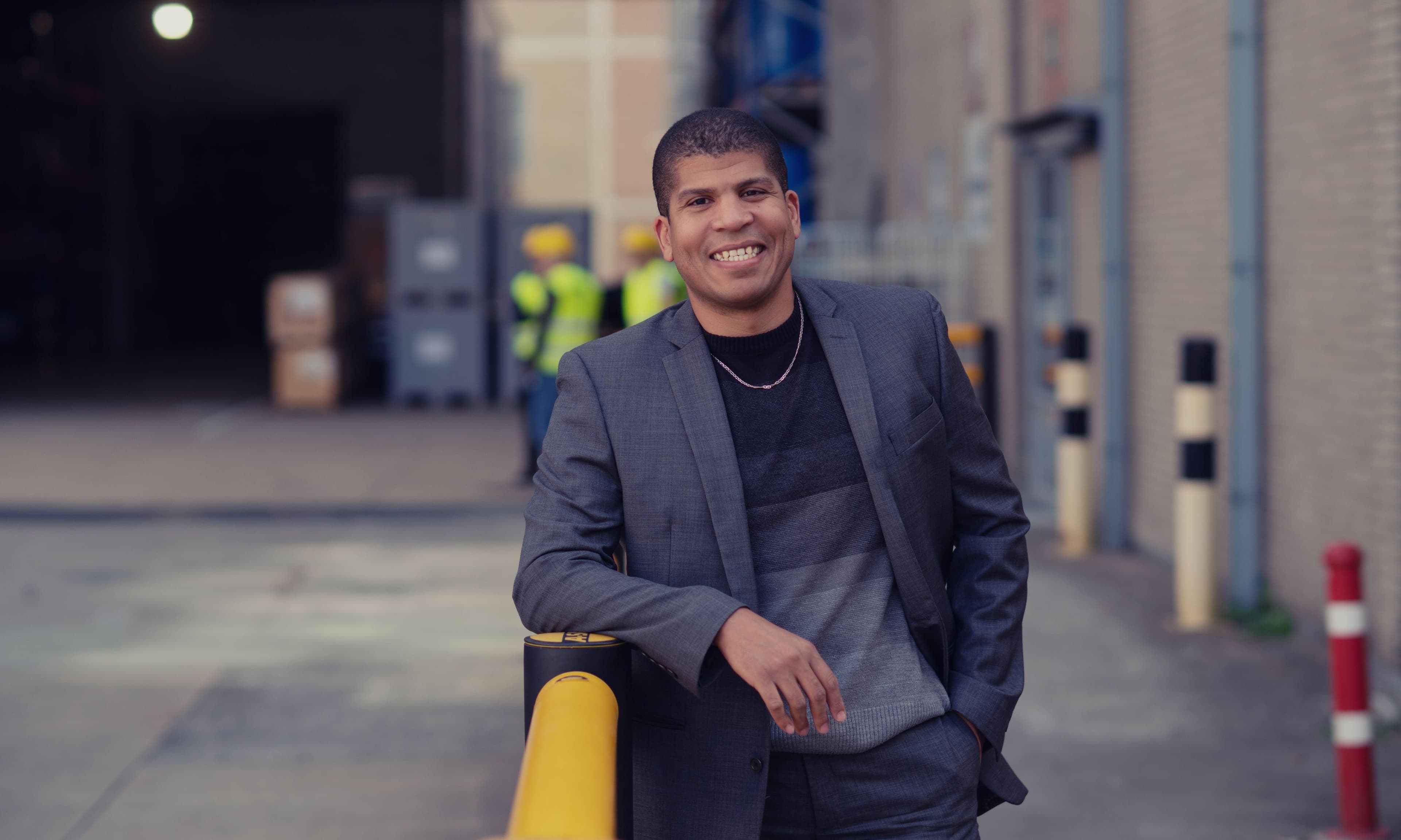 Cesar%20Javier%20Lockhart%20de%20la%20Rosa Smiling man in a gray blazer leaning on a yellow bollard outside an industrial building, with workers blurred in the background.