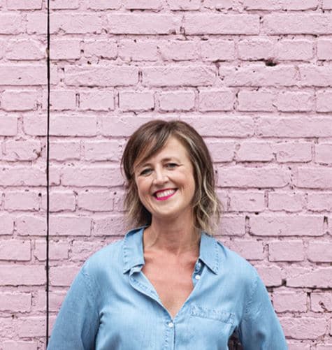 Charlotte%20D%27Hulst Charlotte D'Hulst standing in front of a pink brick wall, smiling in a light blue shirt