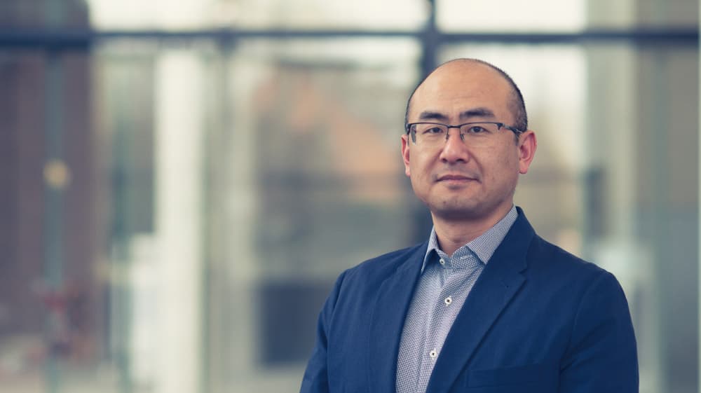 Chengxun-Liu Portrait of Chengxun Liu in a blazer and glasses, standing indoors against a blurred glass background