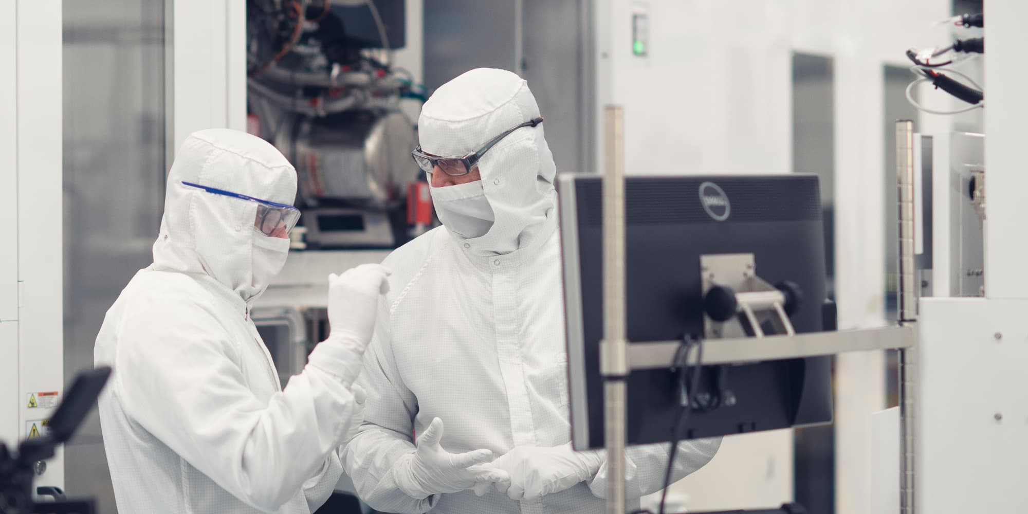 Compute-technology-centers Two cleanroom researchers in protective suits talk beside semiconductor equipment and a monitor