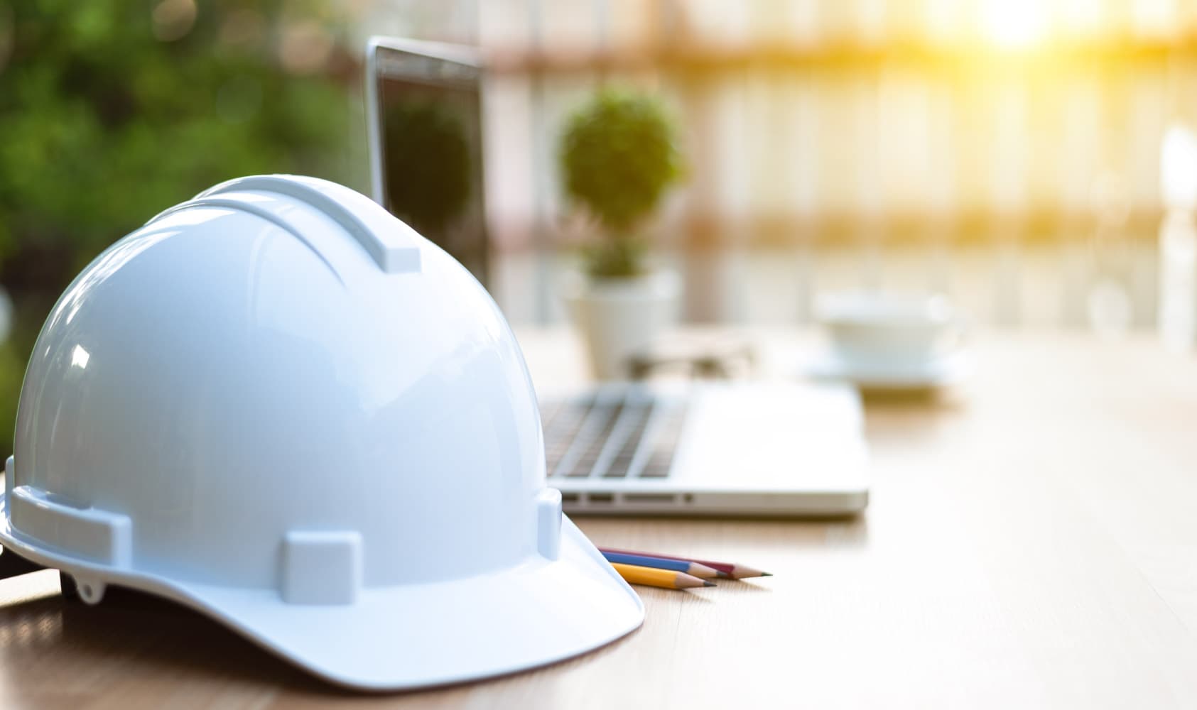 contractor-portal_1 White hard hat on a desk beside a laptop and pencils, with warm sunlight in the background