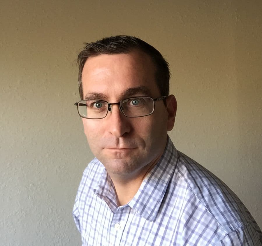 Dan_Rishavy_1 Headshot of Dan Rishavy wearing glasses and a checkered shirt against a plain background