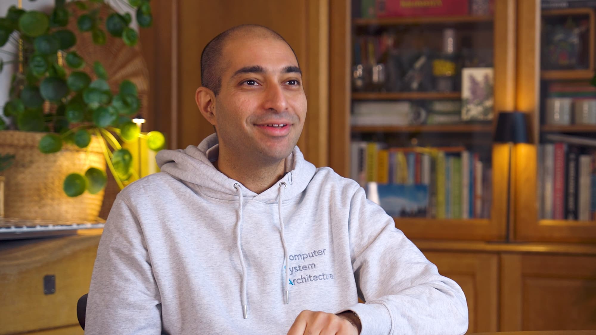 Darayus-Adil-Patel Lachende man in een grijze hoodie, zittend binnen voor boekenplanken en een lamp