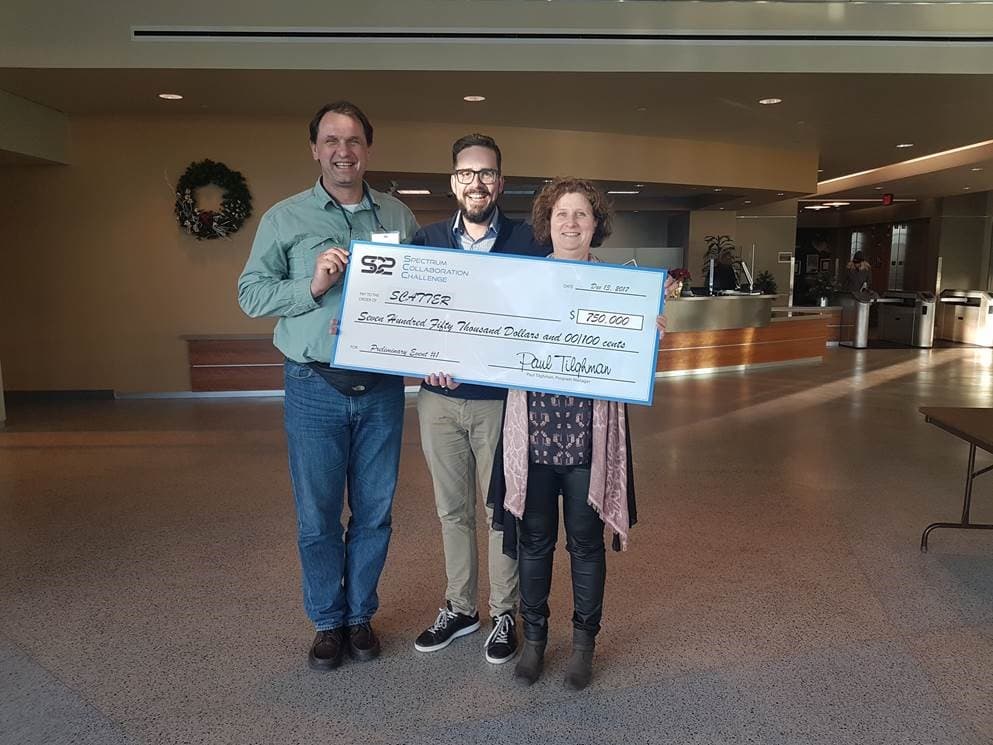 DARPA_1 Three people hold a large $750,000 check in a lobby after a Spectrum Collaboration Challenge award