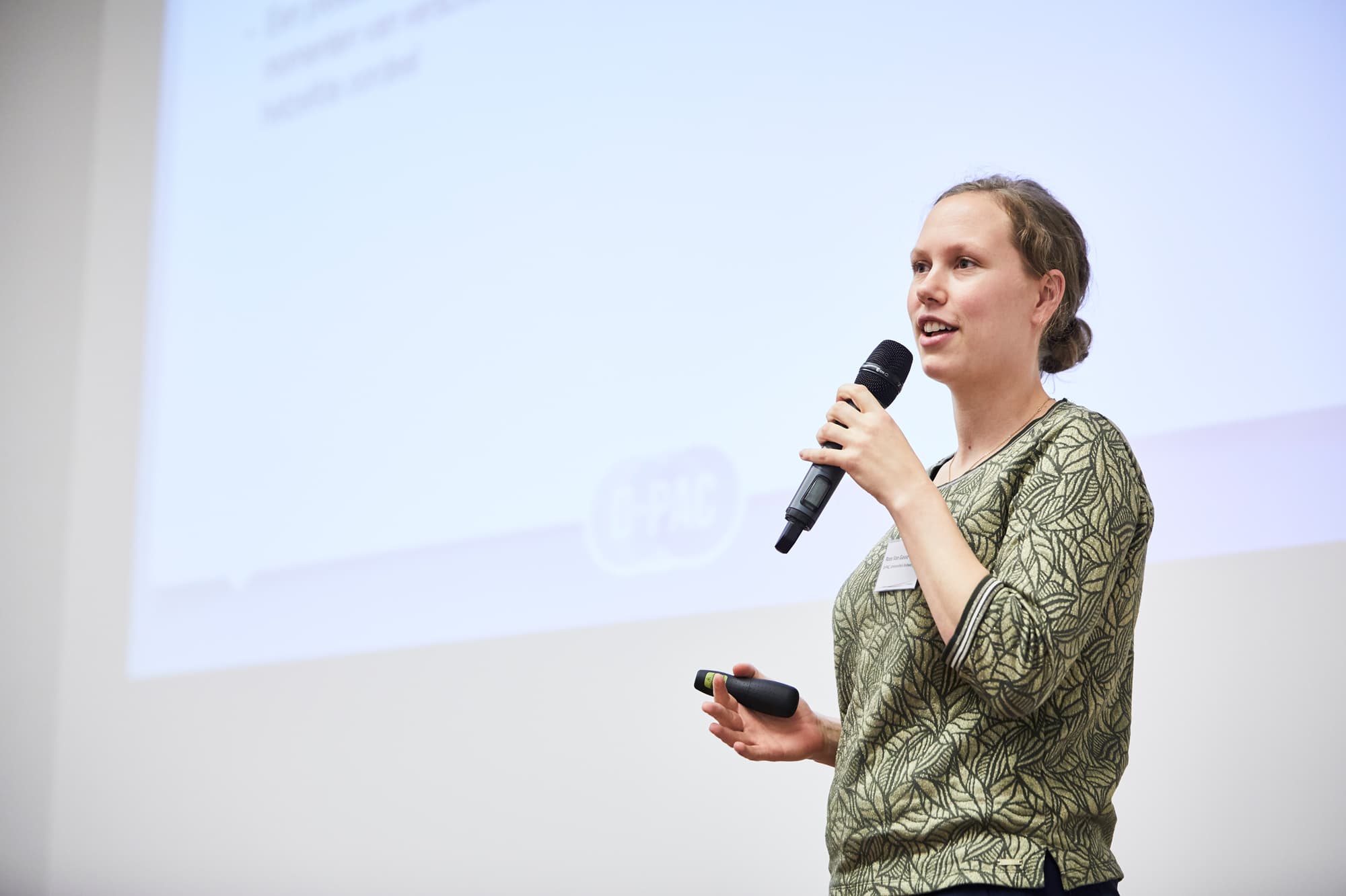 David-Jagersma_Live-Magazines-D-PAC62_1 Woman speaking on a stage with a microphone and presentation screen behind her