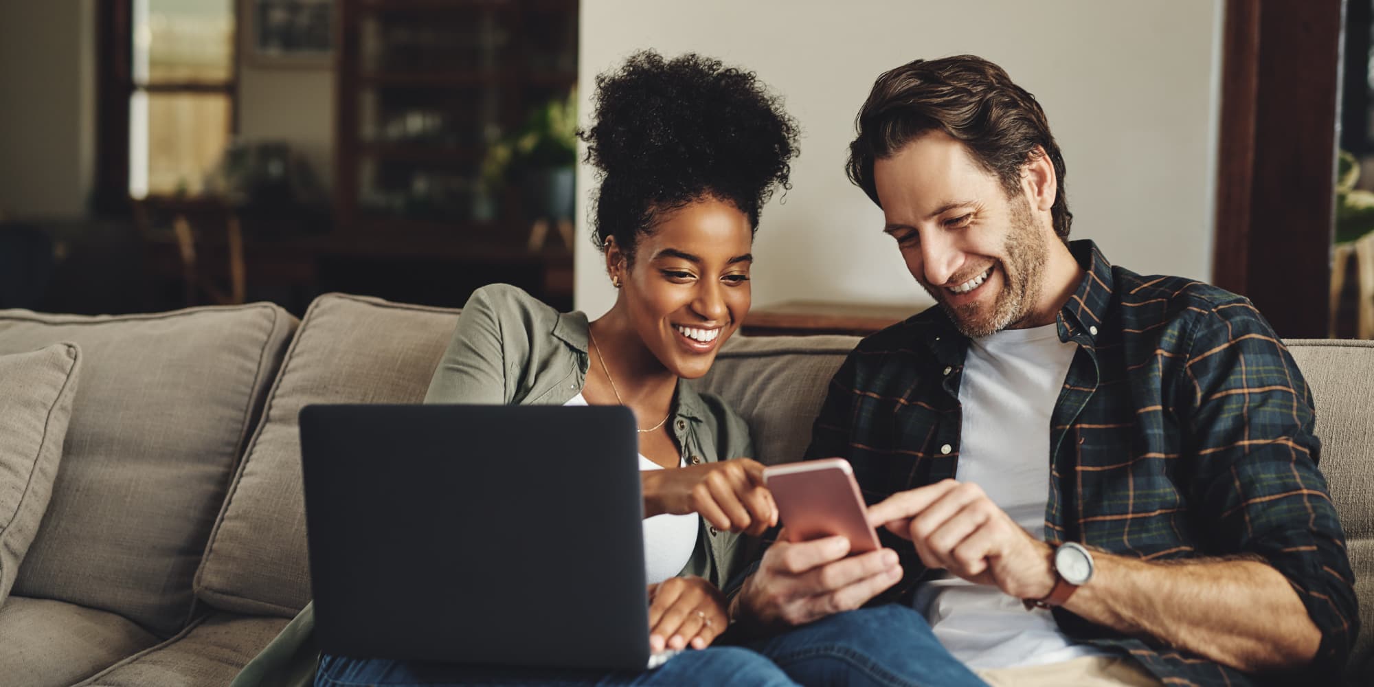 Digimeter-2021 Twee mensen glimlachen naar een smartphone terwijl ze op een sofa zitten met een open laptop naast hen