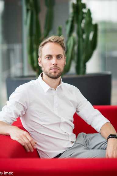 Dries_cr_1 Dries Braeken seated on a red sofa in a white shirt, with tall green plants in the background.