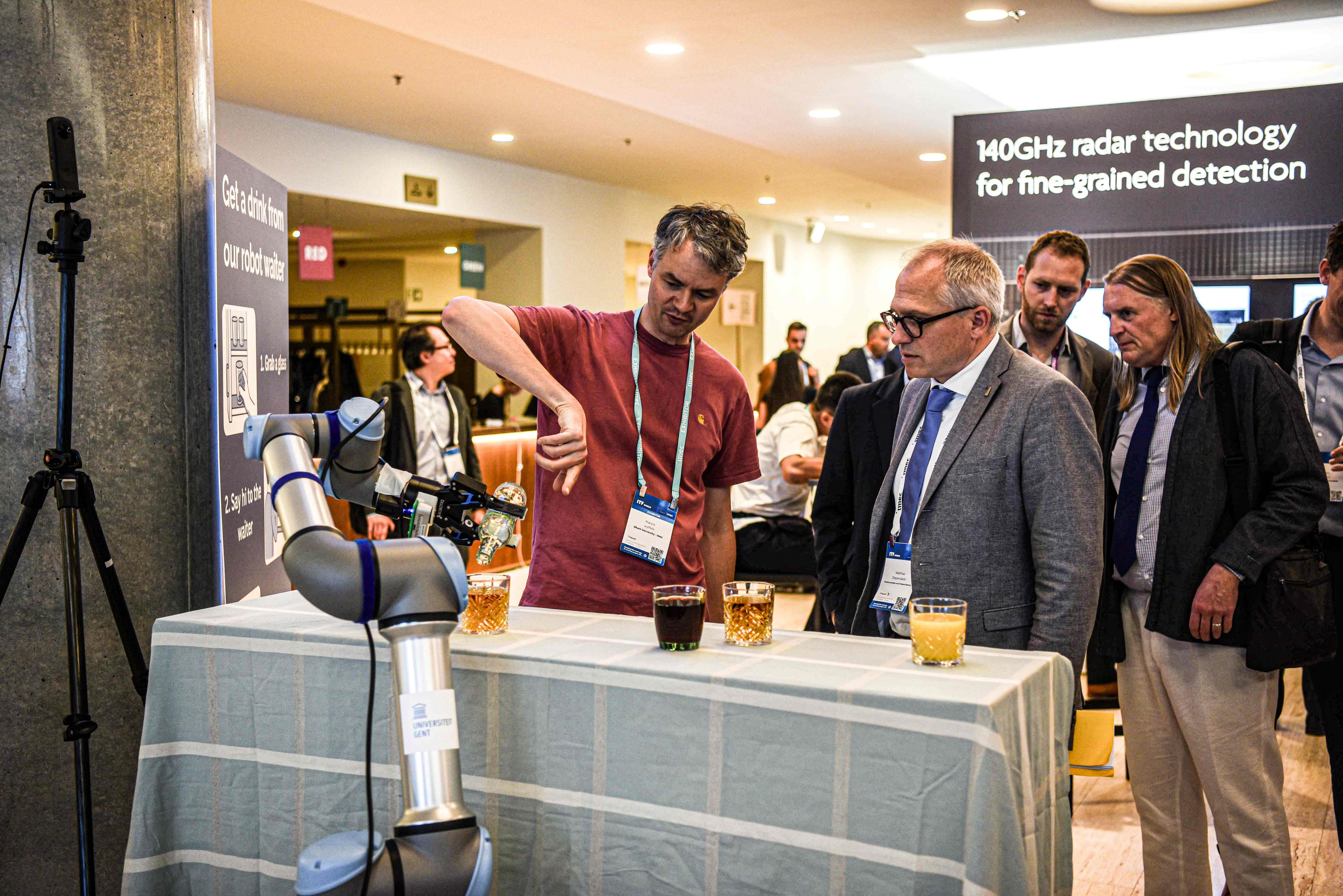 DSC_8926 Een man demonstreert een robotarm op een evenement terwijl bezoekers toekijken, met op de achtergrond een radartechnologiedisplay.