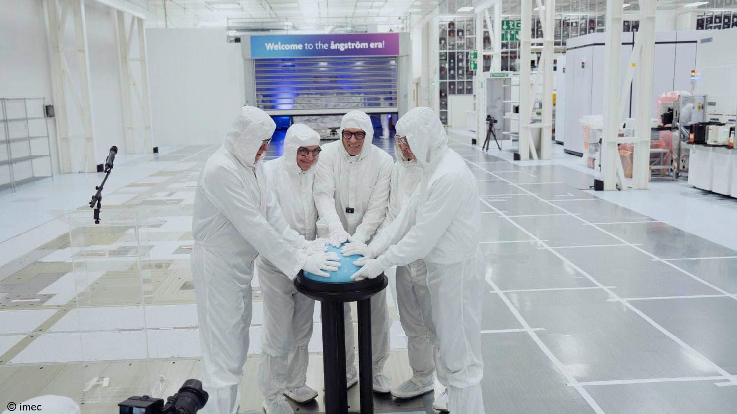 Four cleanroom workers placing hands on blue sphere in imec cleanroom Four cleanroom workers in white suits place their hands on a blue sphere in imec’s cleanroom, with an "ångström era" sign behind them.