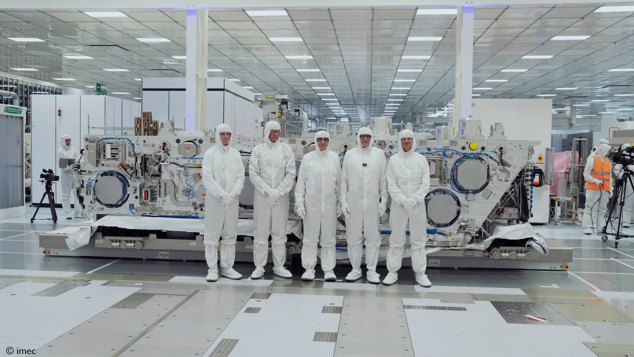 Researchers beside High NA EUV system in semiconductor lab Onderzoekers in cleanroompakken staan naast een High NA EUV-systeem in een halfgeleiderlaboratorium.