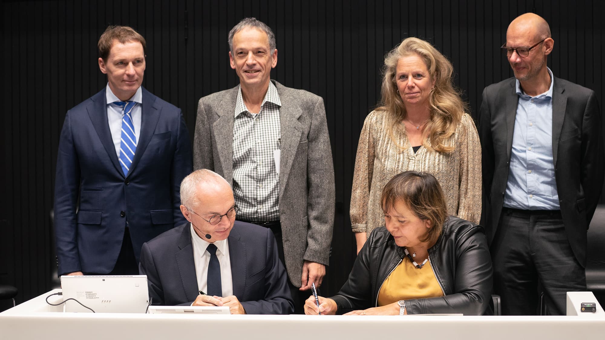 DSC08811 GSK and imec representatives signing an agreement at a table, with colleagues standing behind them.