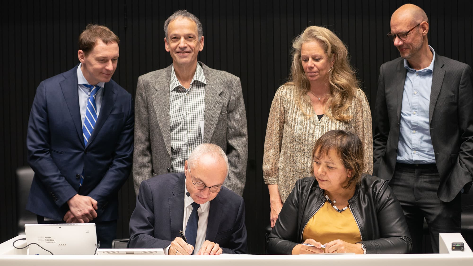 GSK and imec collaboration agreement signing GSK and imec representatives signing a collaboration agreement at a table, with colleagues standing behind them