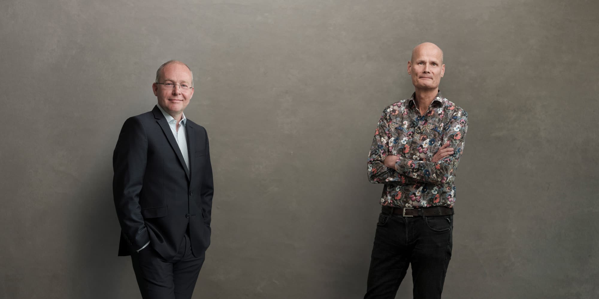 Duo-portret-sywert-brongersma-Nico-Van-Meeteren Two men posing in front of a plain gray backdrop, one in a suit and one in a floral shirt