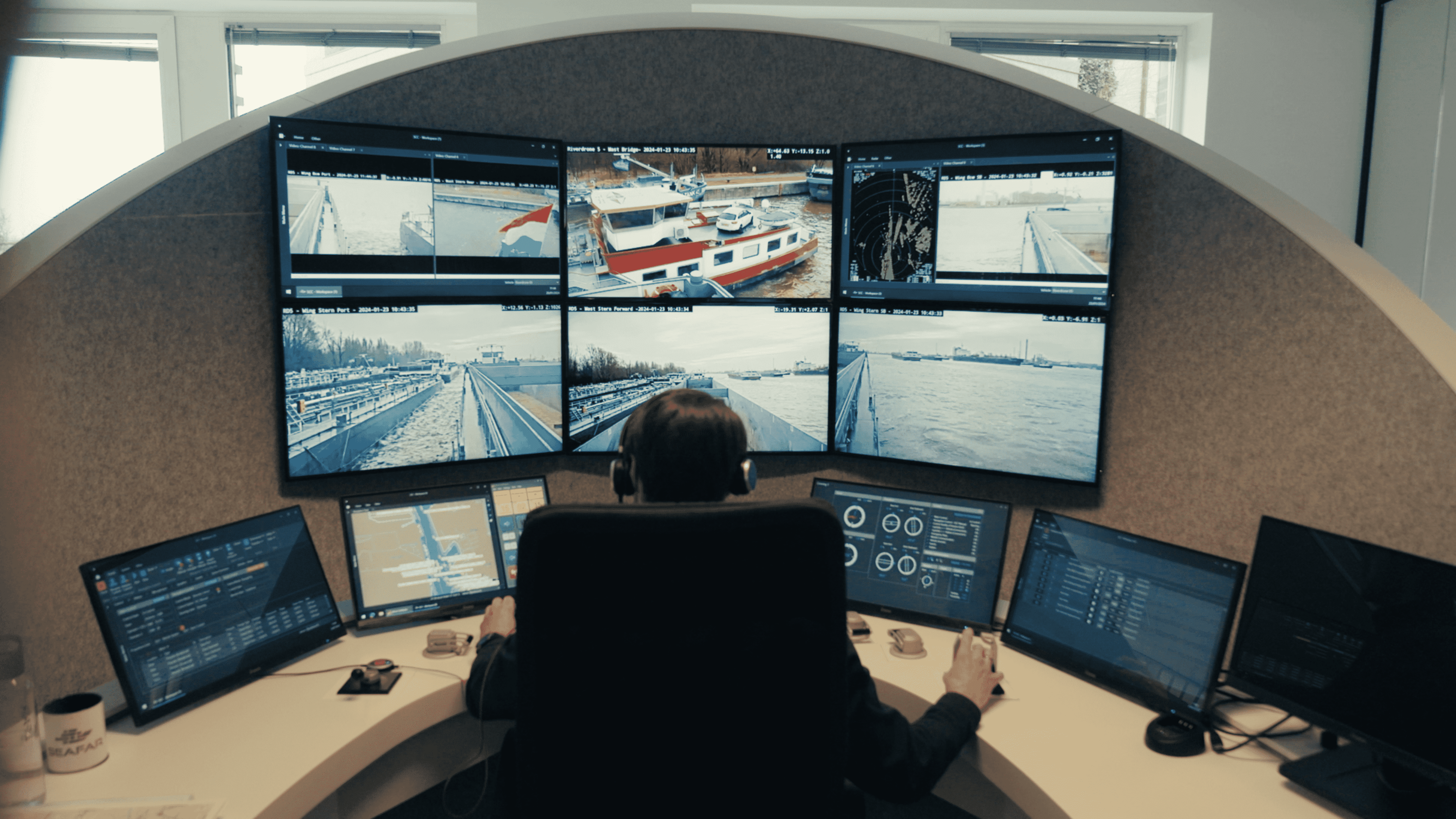Operator monitoring and steering cargo vessel remotely Operator at a control console monitors multiple screens while remotely steering a cargo vessel through a port.