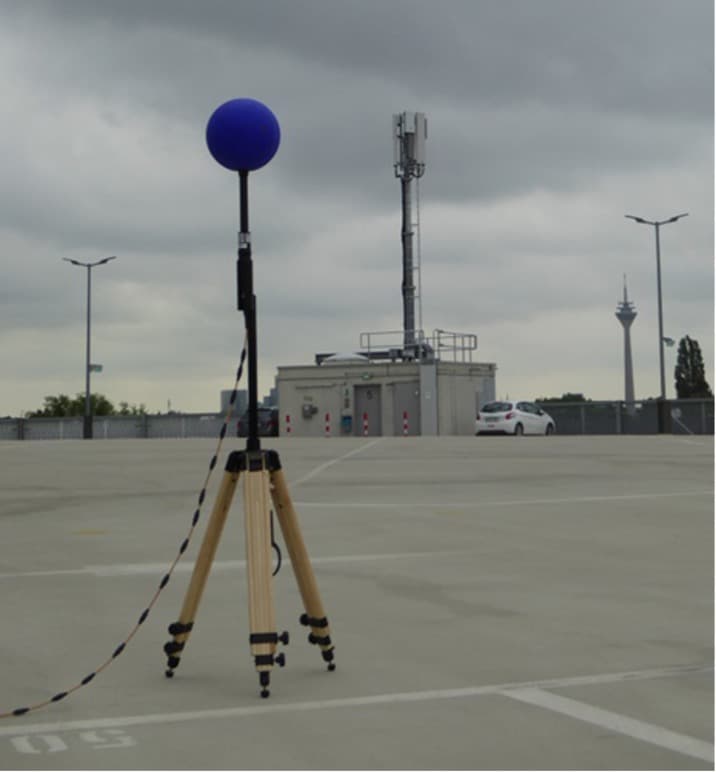 EMF-measurement Blue spherical EMF measurement sensor on a tripod in a parking lot, with a 5G antenna mast in the background.