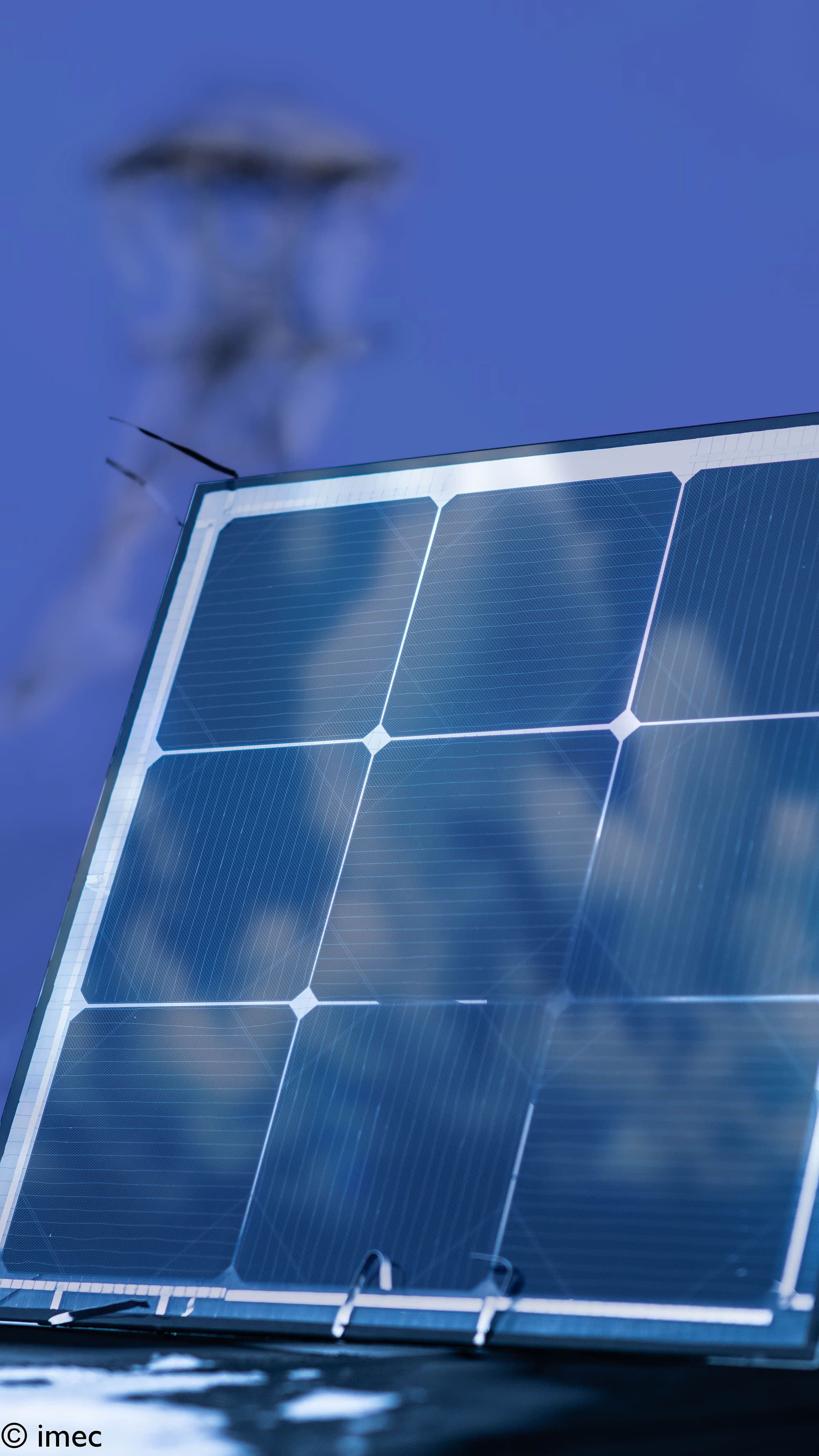 energy-yield-model-bifacial-solarcells002_1 Close-up of a bifacial solar panel against a blue sky, with a blurred wind turbine in the background.