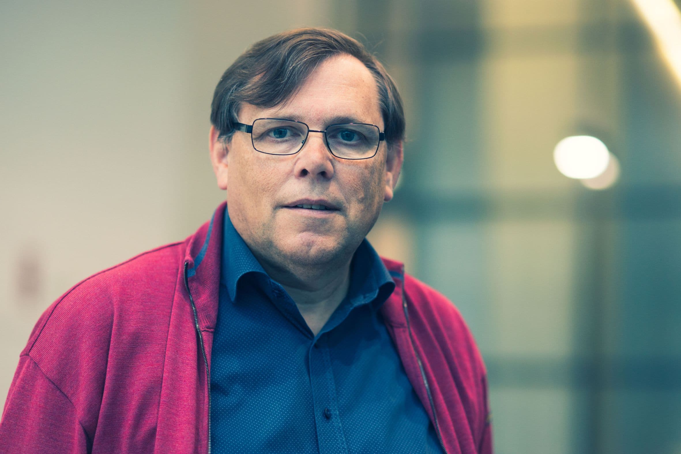 eric_beyne_lr_1 Portrait of Eric Beyne wearing glasses and a red jacket against a blurred indoor background.