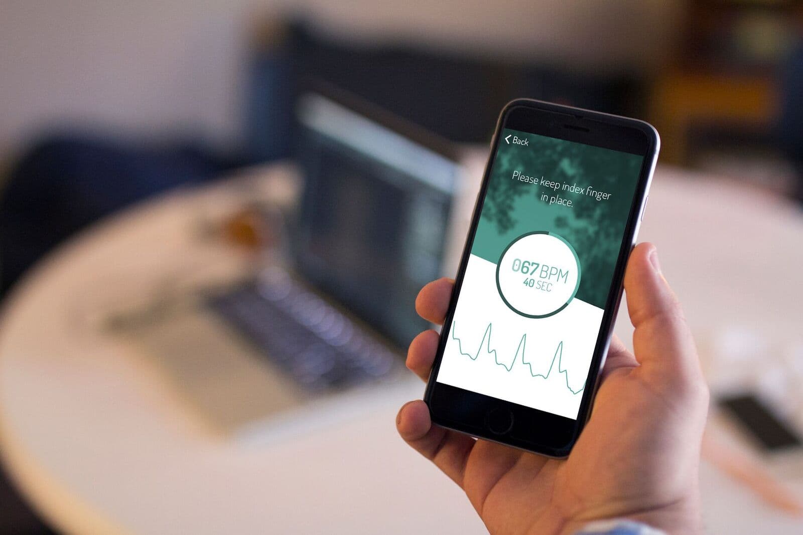 fibricheck Hand holding a smartphone that measures heart rate with a finger on the camera, showing 67 BPM and a pulse graph.