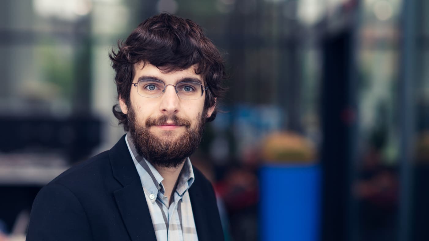Florian001_LR_1 Portrait of Florian De Roose wearing glasses and a dark blazer, with a blurred indoor background