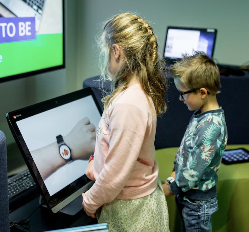 Children looking at smartwatch touchscreen in classroom Two children look at a touchscreen showing a smartwatch on a wrist in a classroom setting